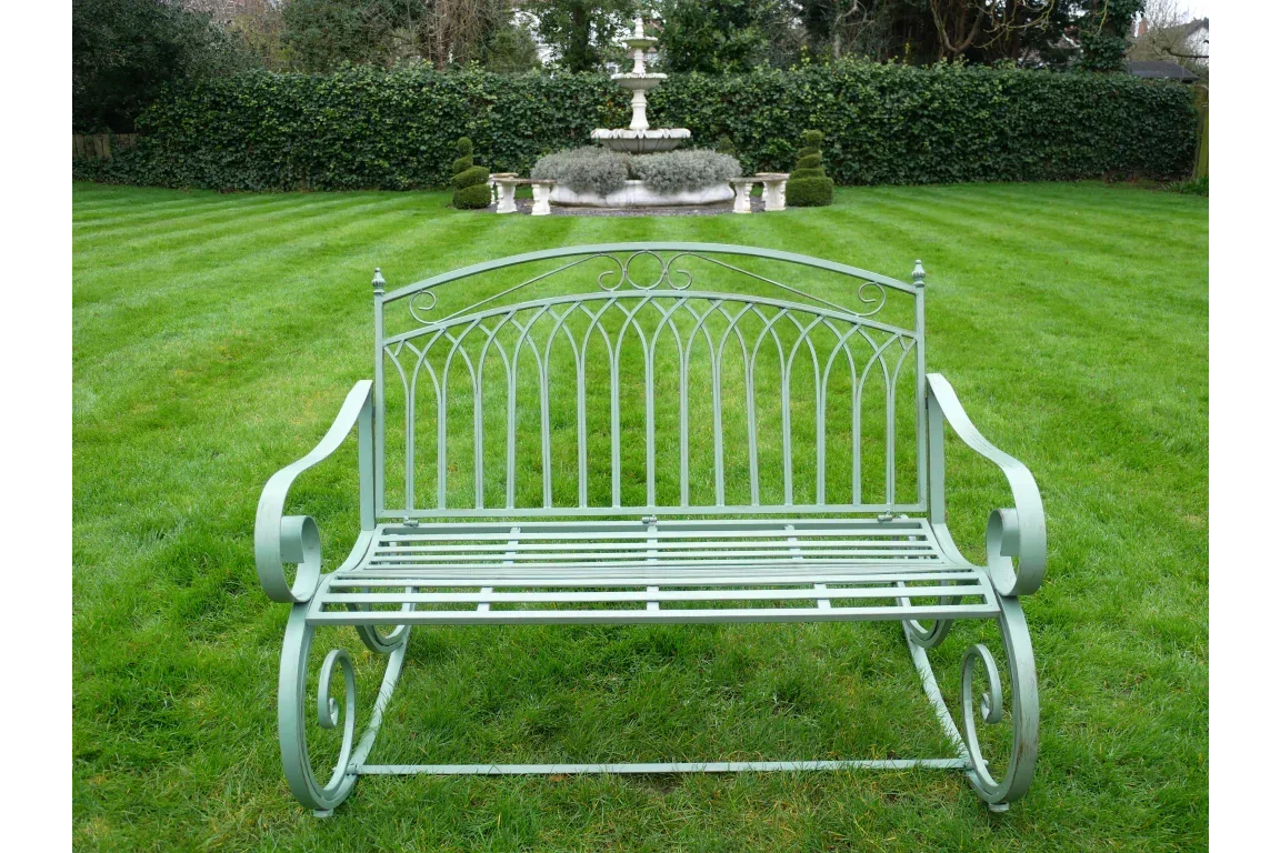 Green metal garden bench with scroll arms on lawn, fountain in background
