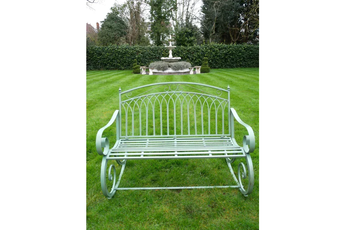 Green metal garden bench with scroll armrests on lawn, fountain in background