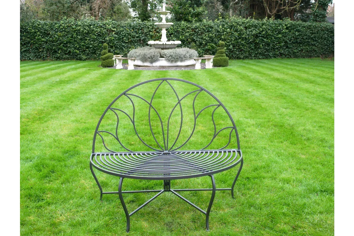 Black metal garden bench with curved backrest on lawn, fountain in background