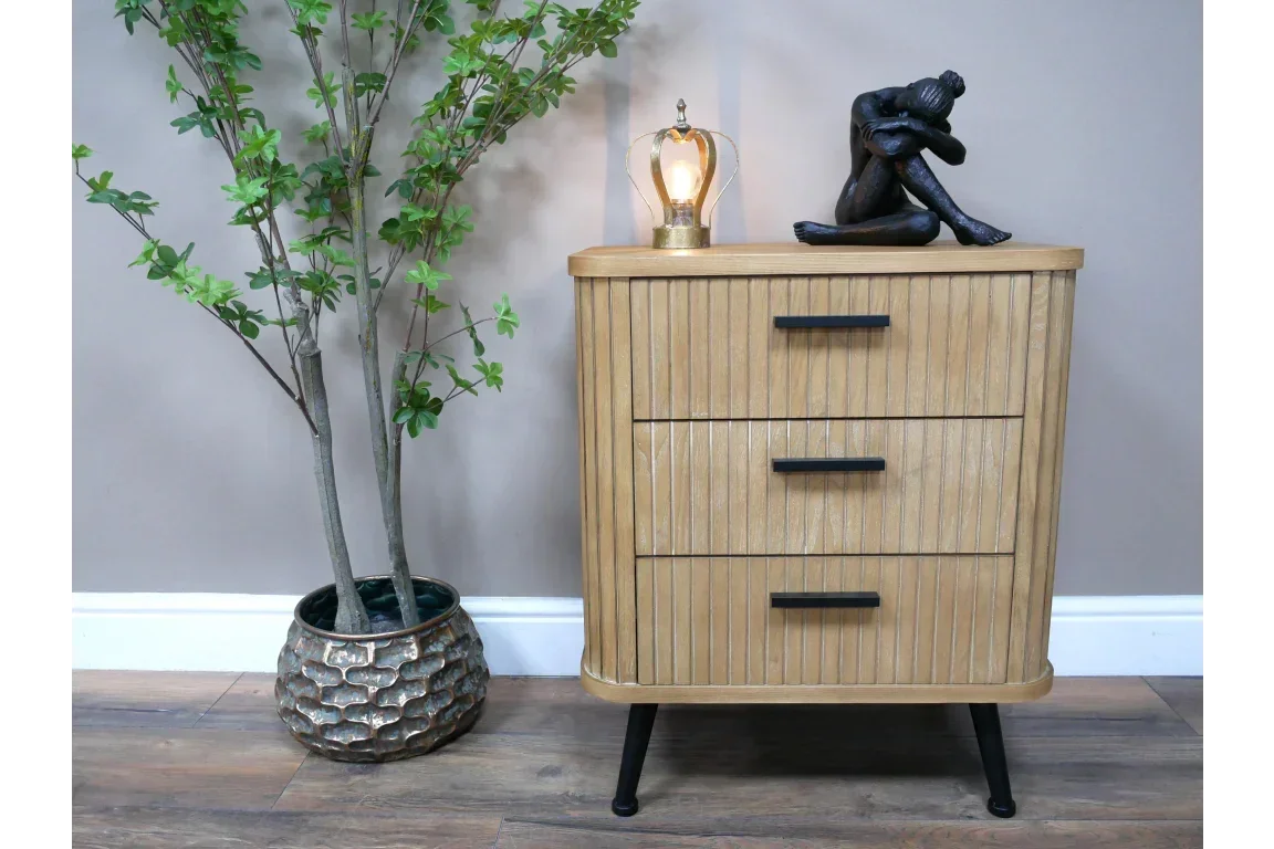 Oak bedside table with three drawers, black handles, lamp, and figurine, next to potted plant