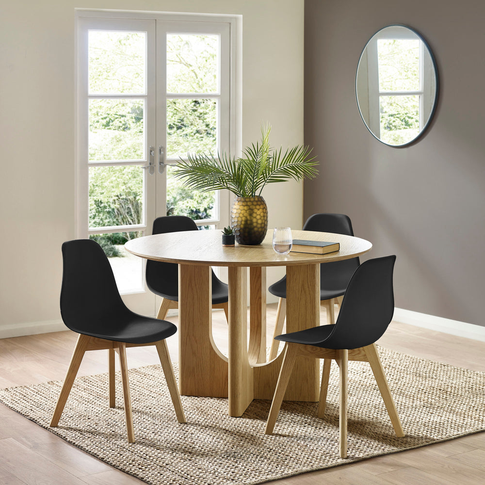 Black chairs with wooden legs around round light oak table in room with glass doors, mirror, plants, and rug