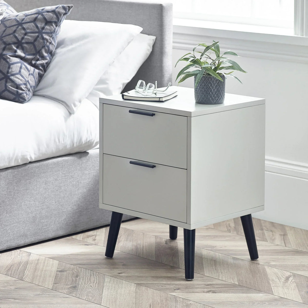 Silk grey two drawer bedside table with black legs beside bed, plant and glasses on top