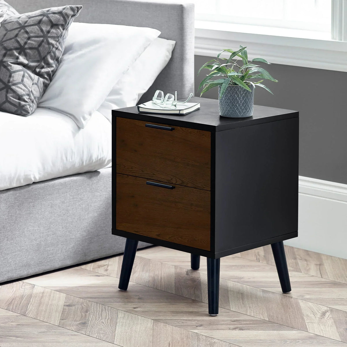 Walnut and black 2 drawer bedside table beside grey bed, plant and book on top