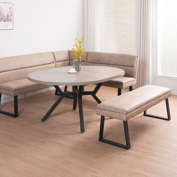 A round dining table with a light gray wood-look top and black metal legs, paired with a beige upholstered bench and matching curved sectional seating in a bright room with wood flooring.