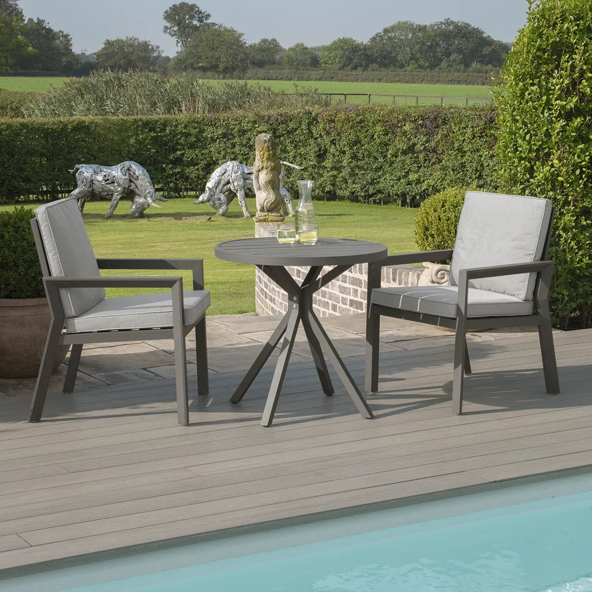 Grey aluminium garden bistro set with round table and two cushioned chairs by a pool