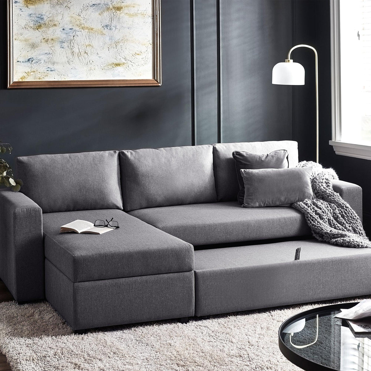Grey sofabed with storage feature in modern living room beside large window and table