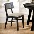 Black dining chair with natural fabric seat and rattan backrest angled left on woven rug beside round black table