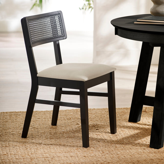 Black dining chair with natural fabric seat and rattan backrest angled left on woven rug beside round black table