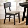 Black dining chair with natural fabric seat and rattan backrest angled left on woven rug beside round black table