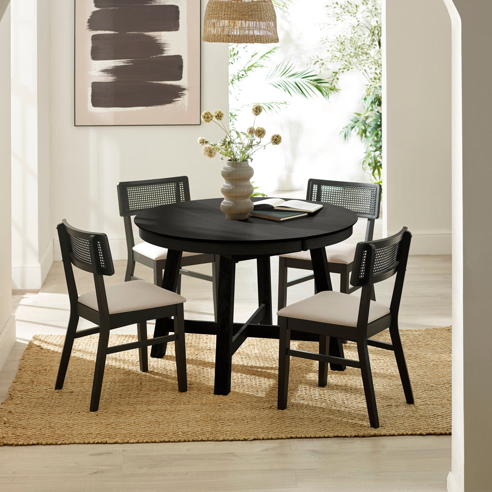 Bright room with round black wood dining table and four black rattan-back chairs with cream seats on woven rug.