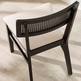 Black wooden dining chair with natural rattan back and cream fabric seat, viewed from back angle on wooden floor.