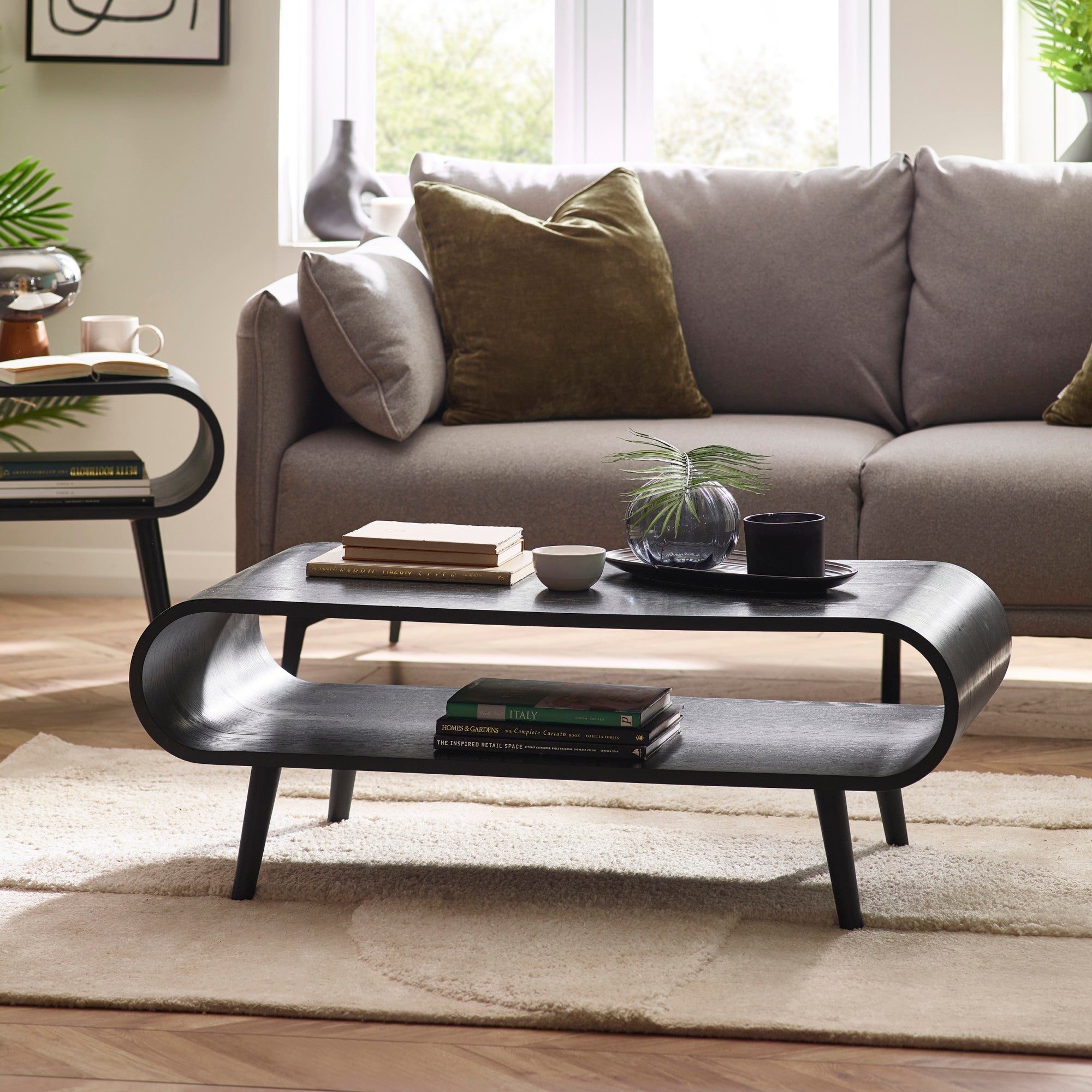 Black wood coffee table with curved ends, lower shelf holding books, placed on beige rug in living room