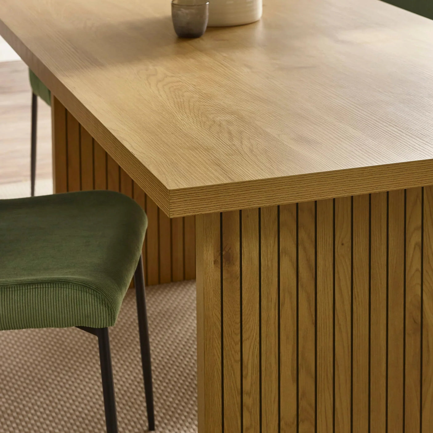 Oak dining table with vertical slat base and green upholstered chair on textured rug