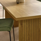 Oak dining table with vertical slat base and green upholstered chair on textured rug