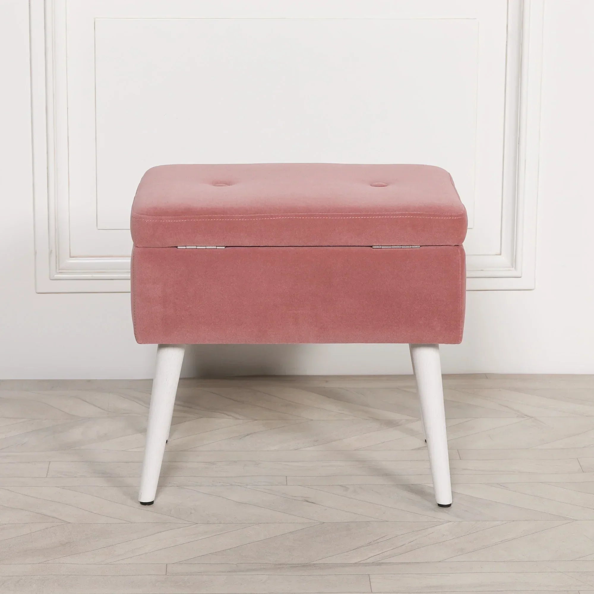 Pink velvet storage ottoman bench with white legs against light wall and wooden floor