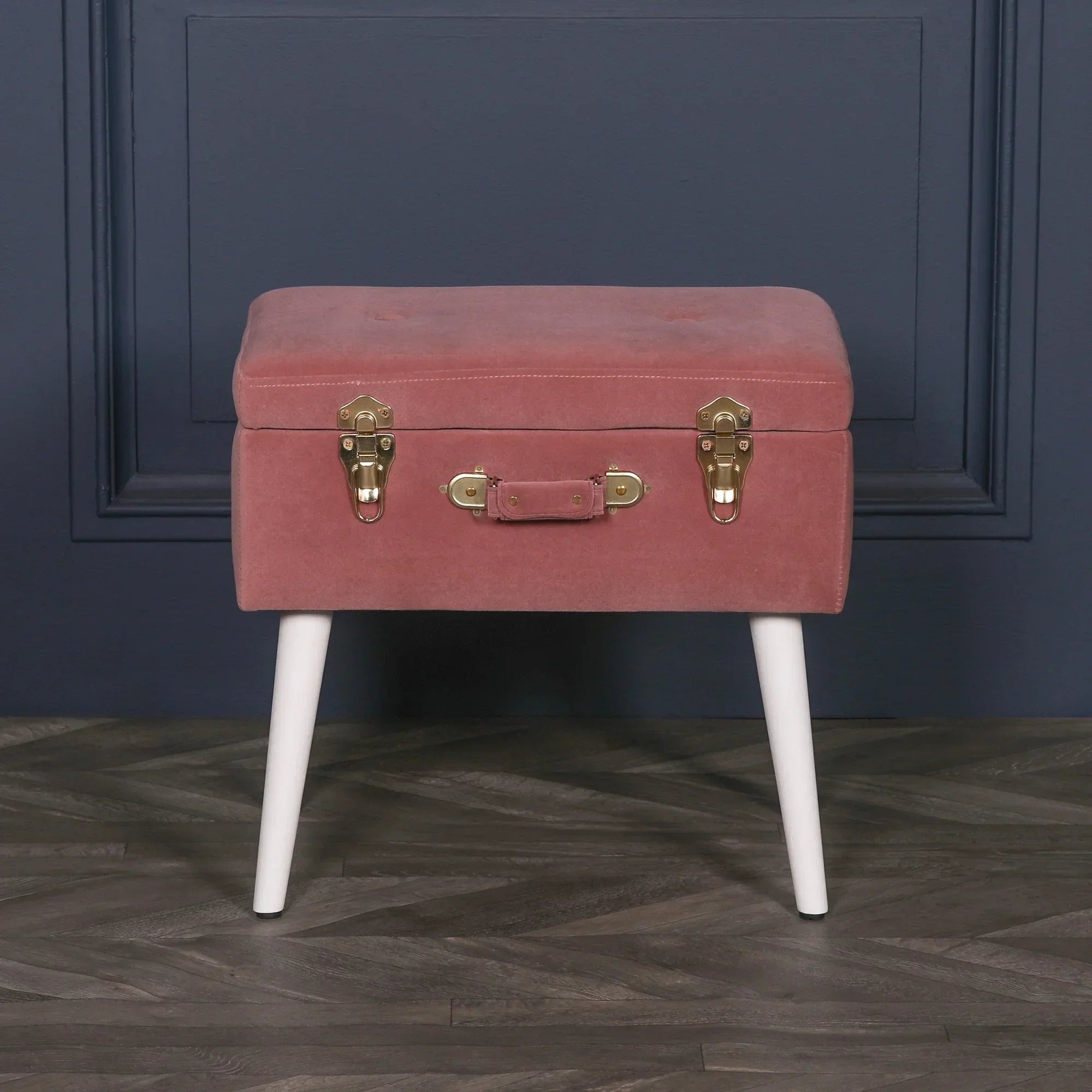Blush pink velvet ottoman with suitcase design and white legs against blue wall