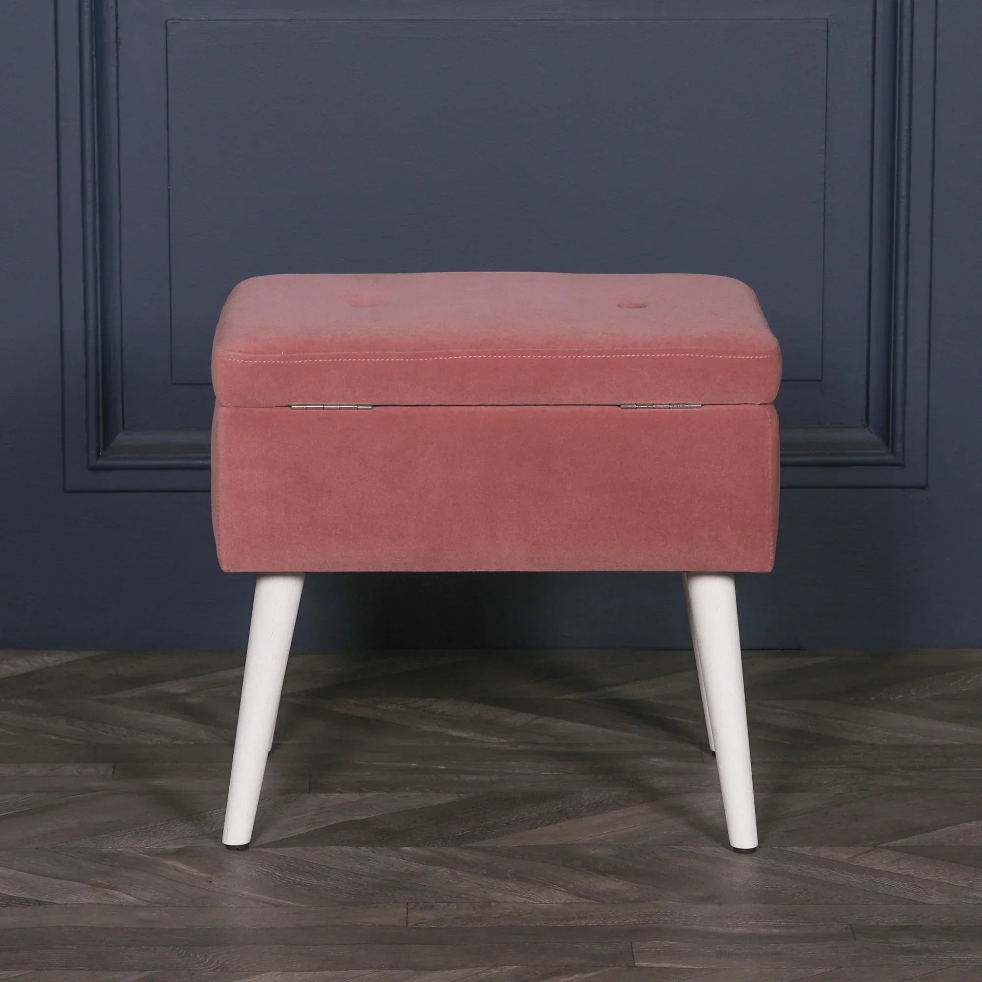 Blush pink velvet storage stool with white angled legs against dark blue wall