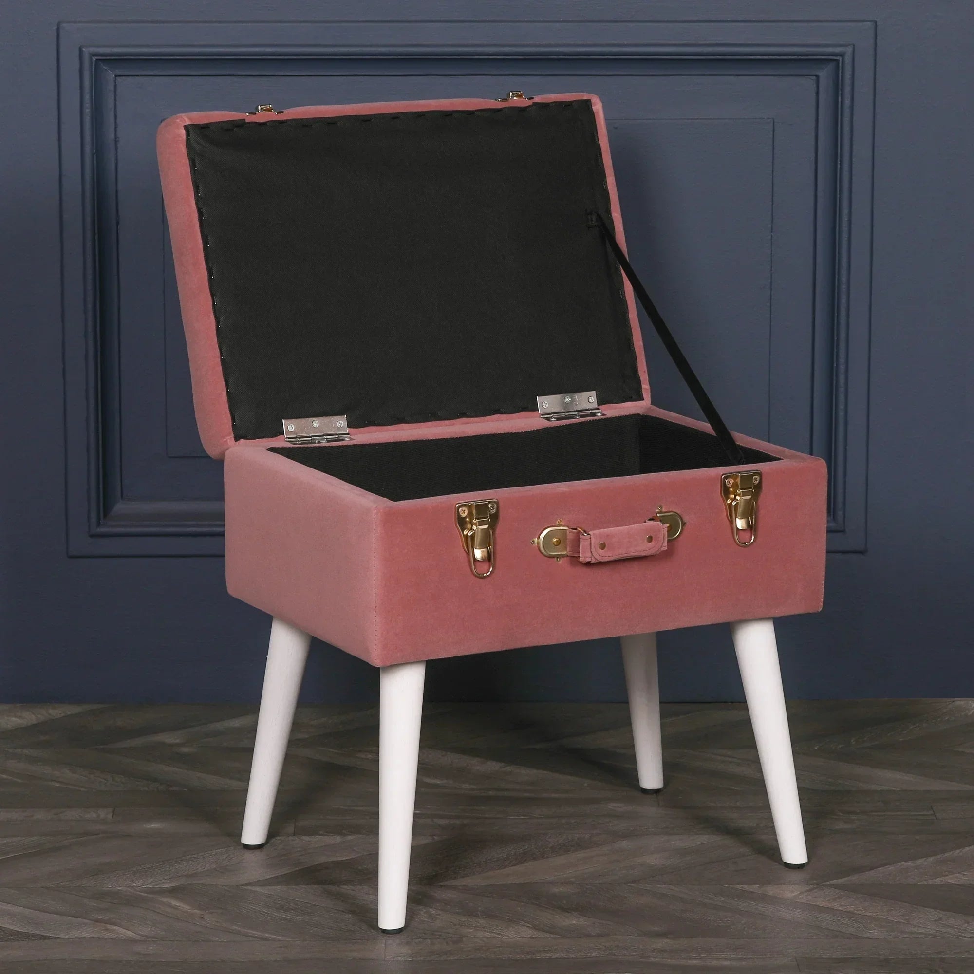 Pink velvet storage ottoman with white legs and open lid against dark blue wall