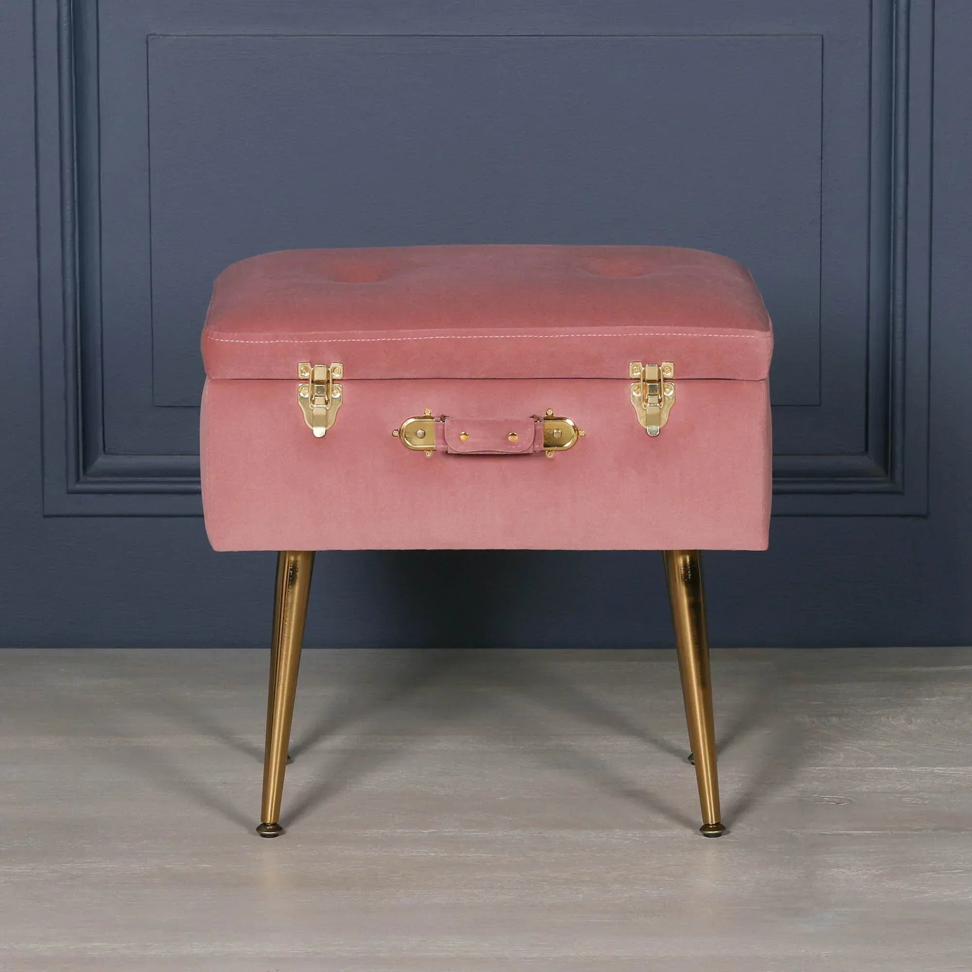Pink velvet ottoman with gold legs and suitcase-style handle against navy wall