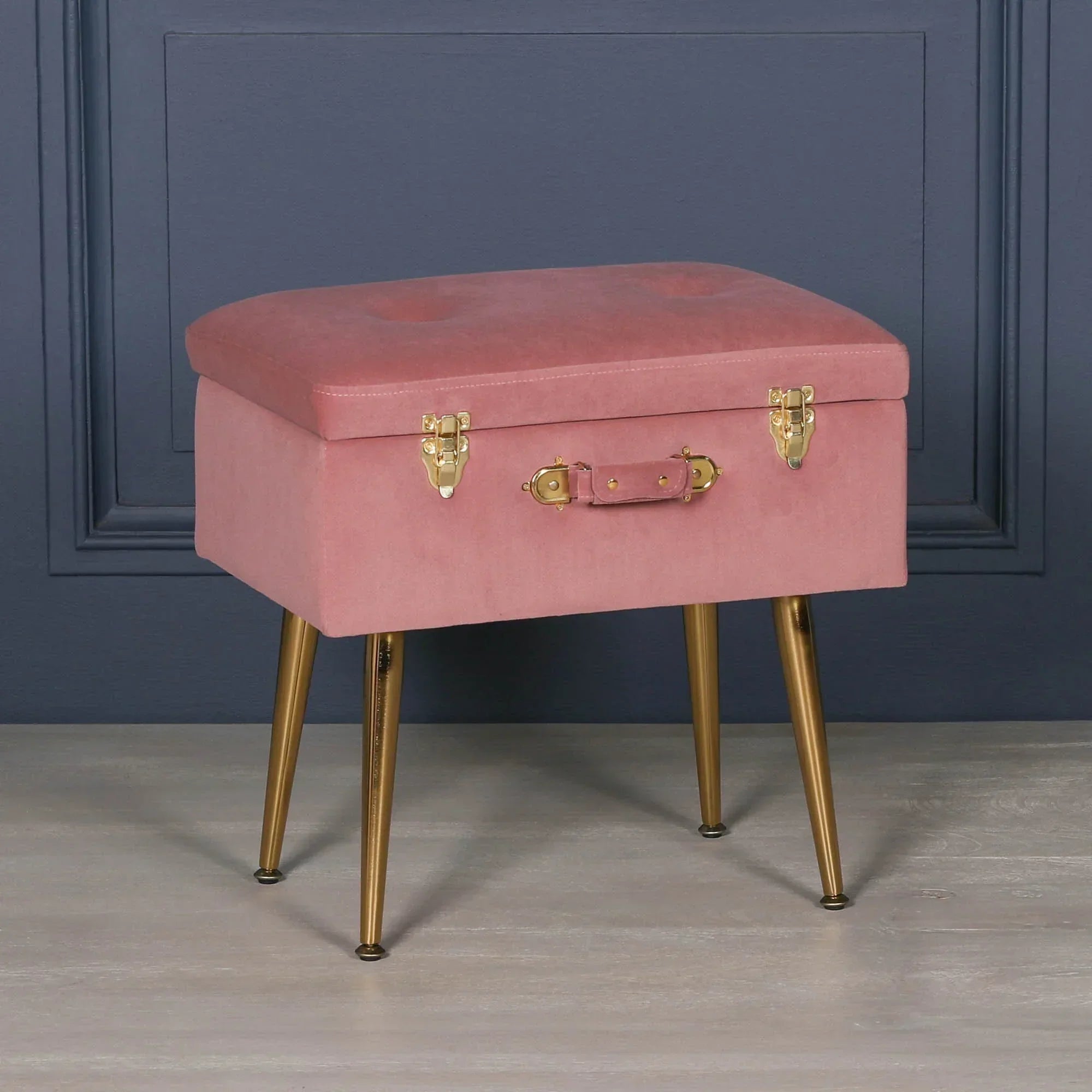 Pink velvet storage ottoman with gold metal legs and brass latches against dark wall