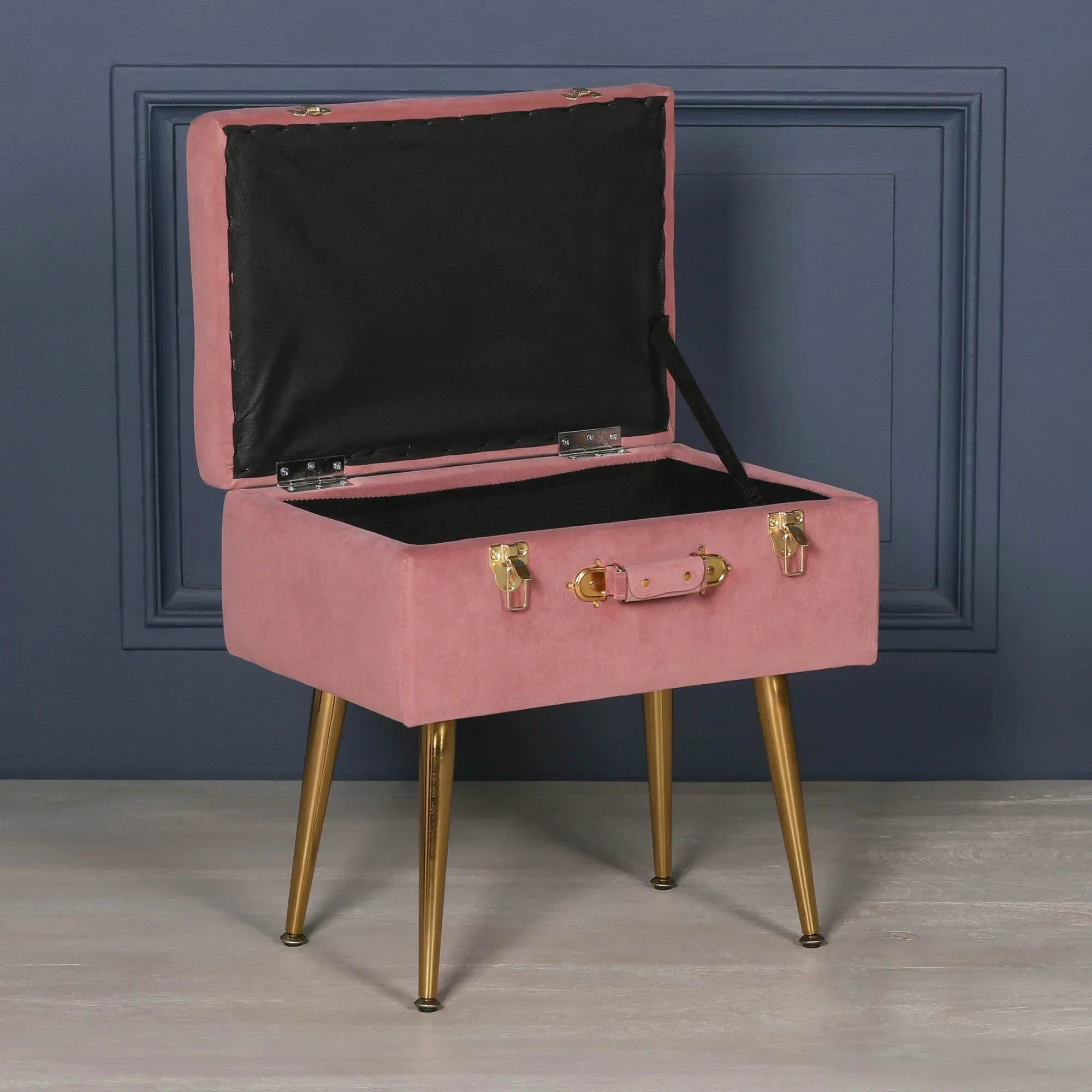 Pink velvet storage stool with gold legs and open lid against dark blue wall