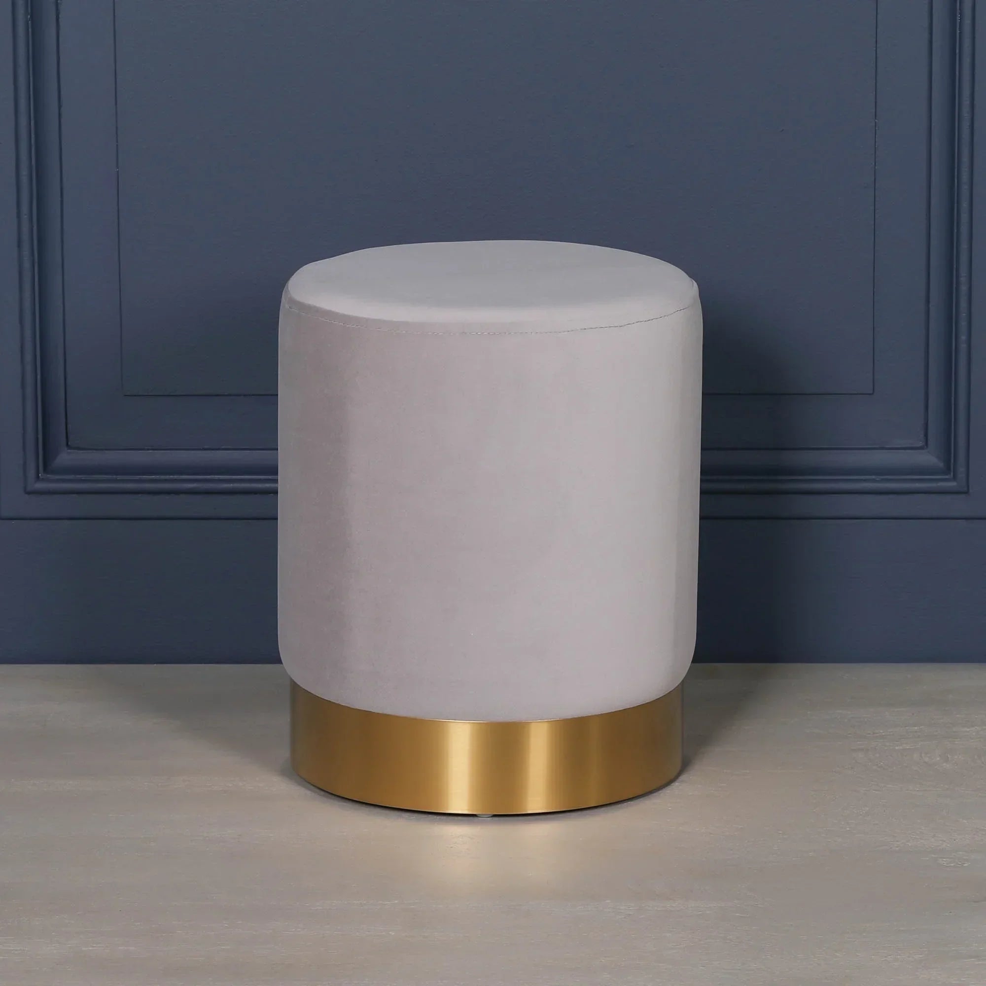 Light grey velvet ottoman stool with gold metal base against blue wall