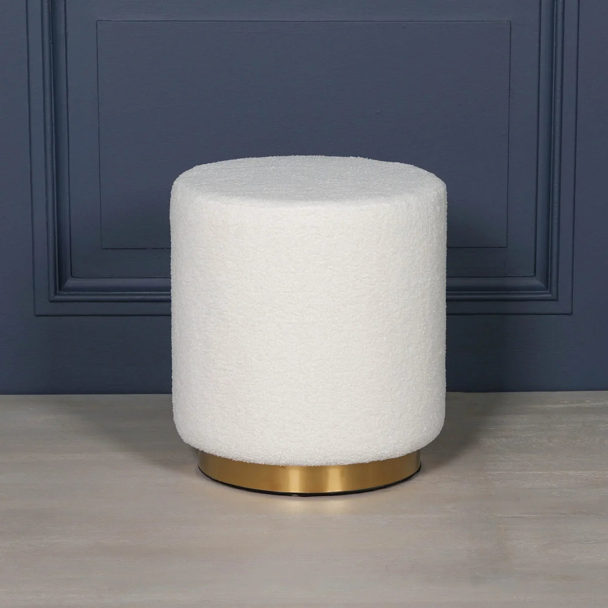 Round white boucle ottoman with gold base on wooden floor against navy wall
