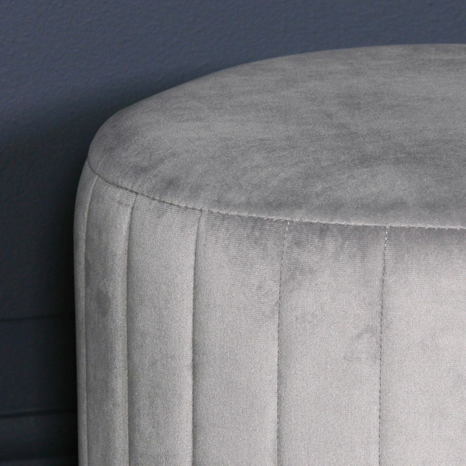 Light grey velvet footstool with vertical stitched panels against dark wall