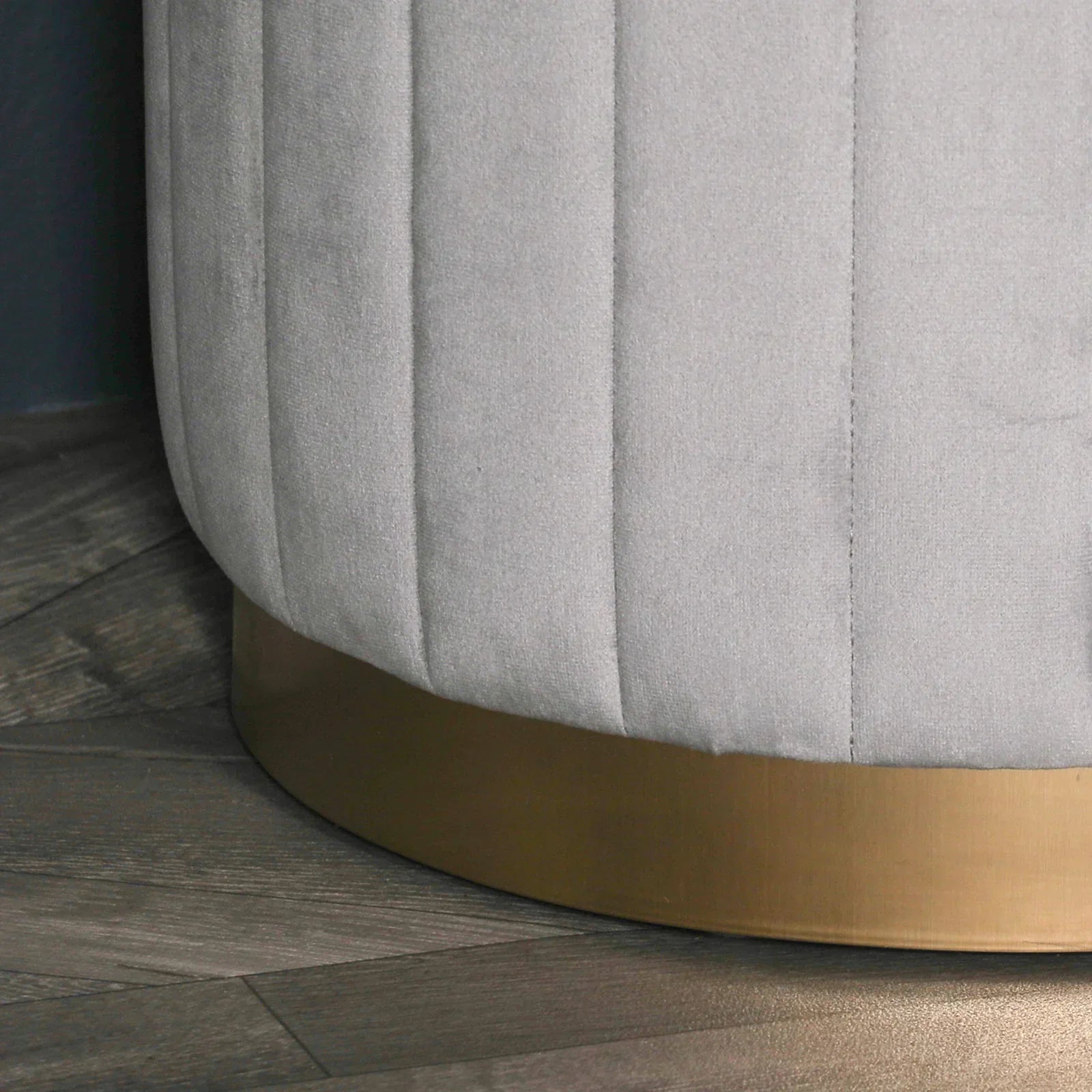 Close view of grey velvet ottoman with vertical stitching and gold metal base on wood floor