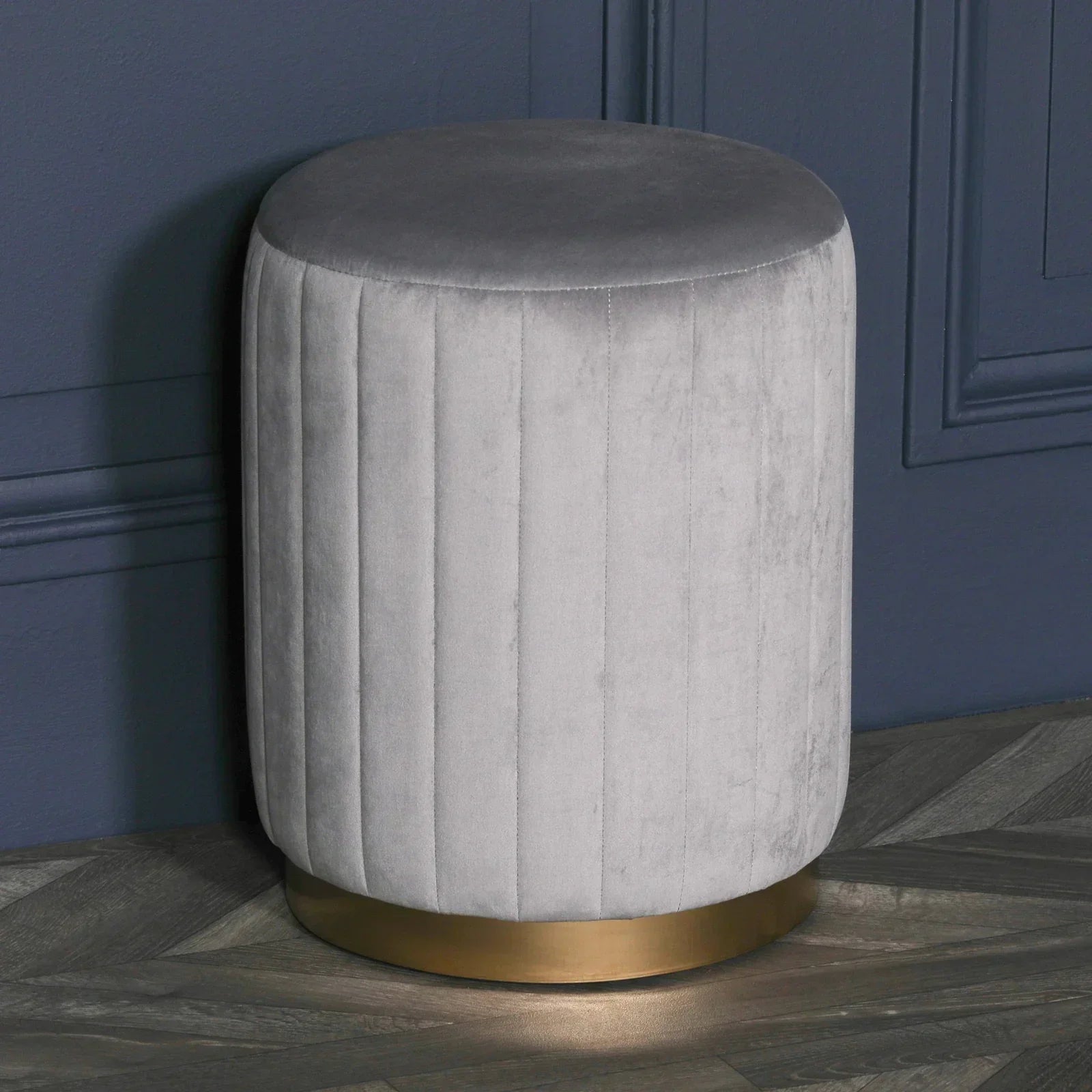 Grey velvet ottoman stool with gold base against dark blue wall on wooden floor
