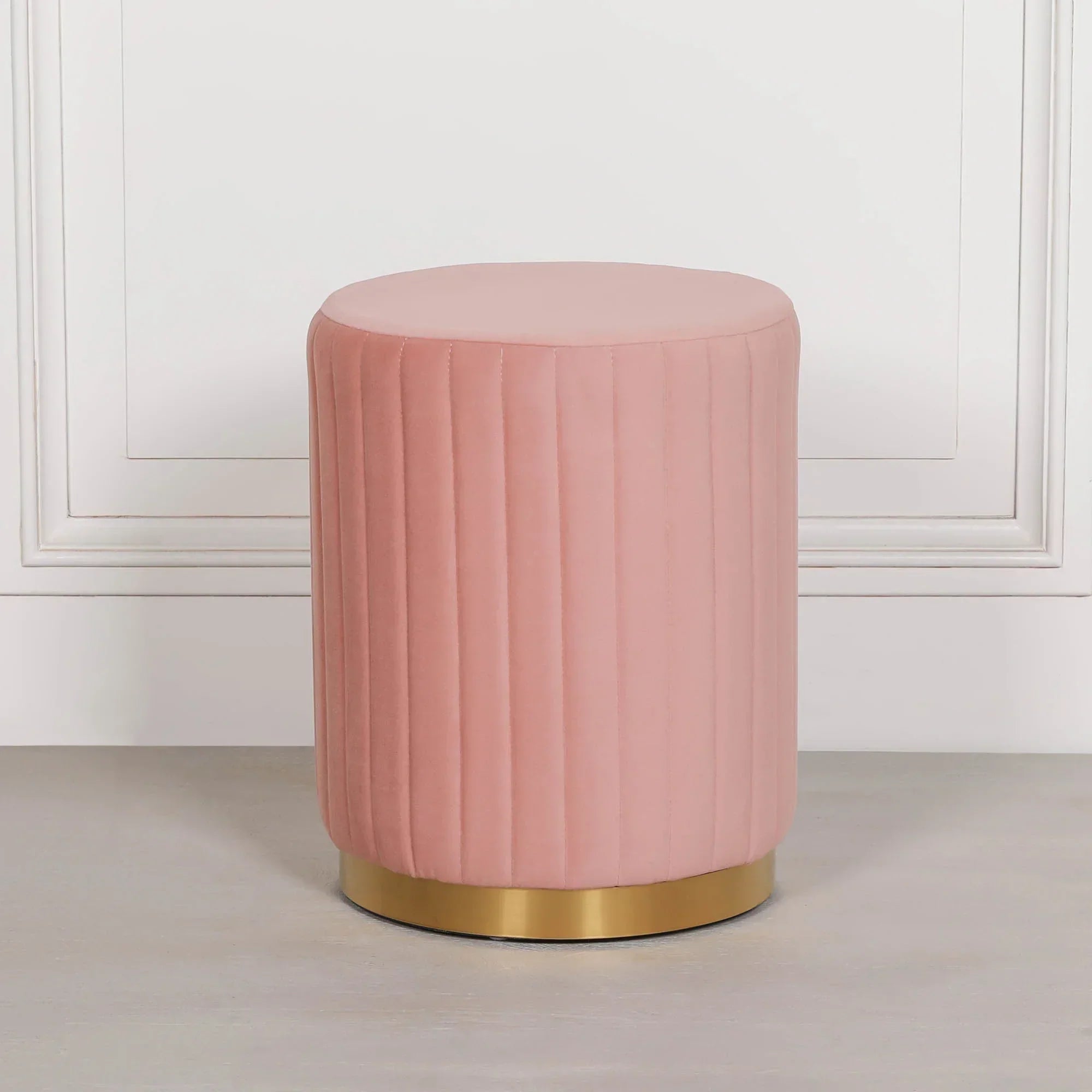 Pink velvet ottoman stool with vertical stitching and gold base against white wall
