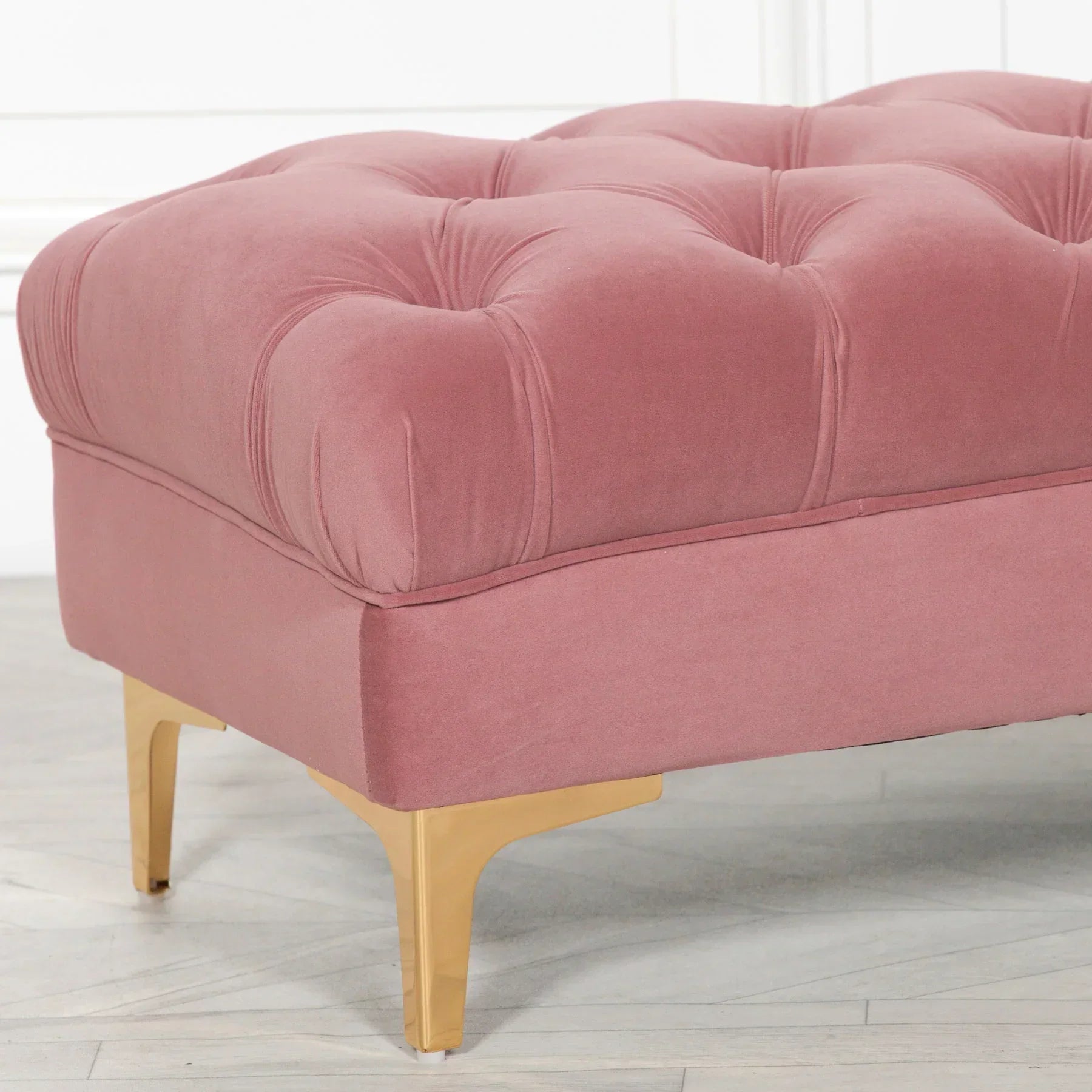 Blush pink velvet ottoman with tufted top and gold metal legs on wood floor