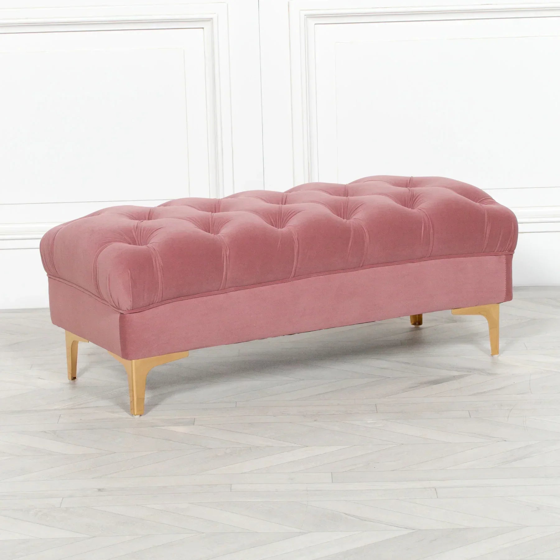 Pink velvet ottoman bench with tufted upholstery and gold metal legs on wooden floor
