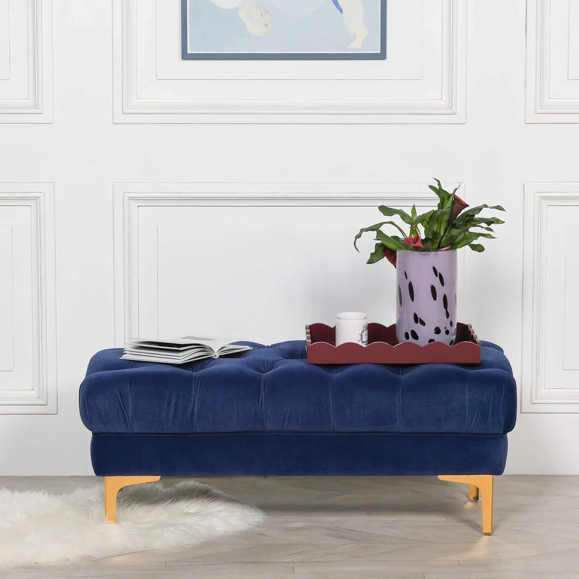 Blue velvet ottoman bench with gold legs, tray, plant and open book against white wall