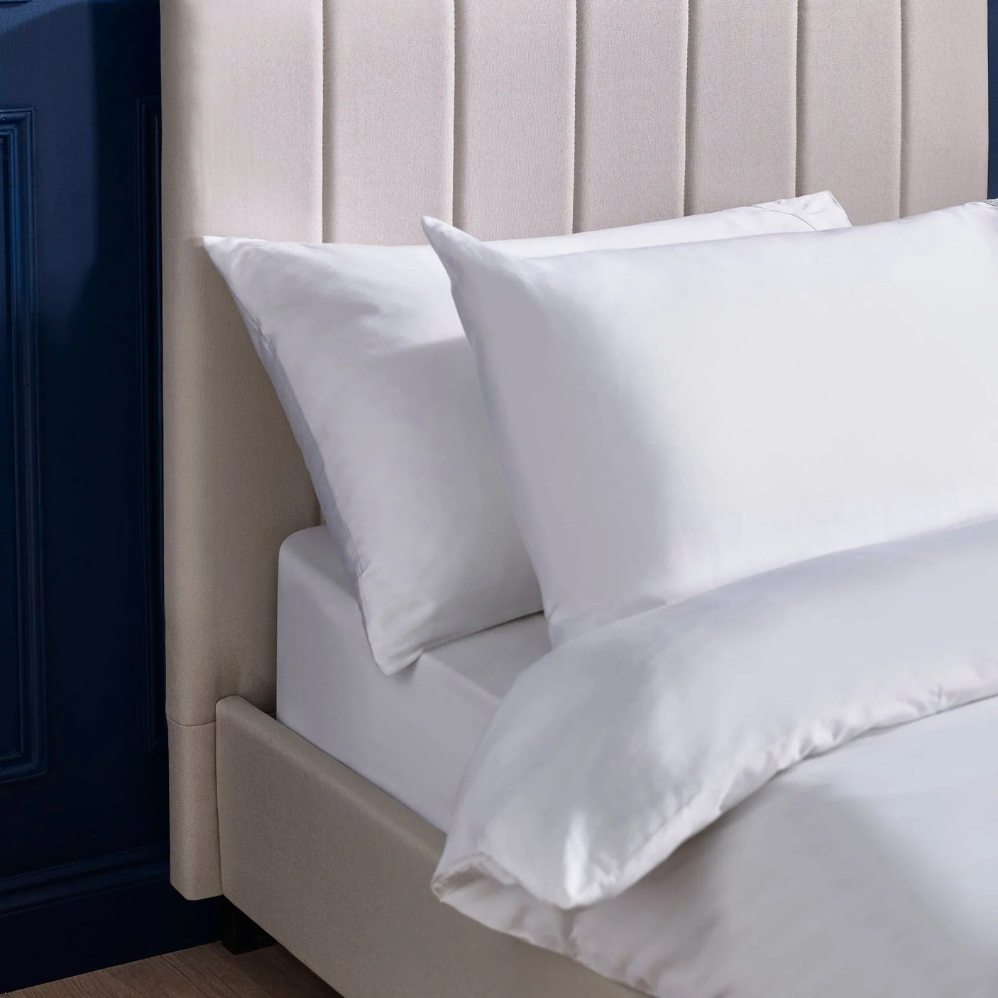 White bedding set with pillowcases on beige upholstered bed against navy wall