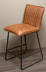Cognac bar stool with quilted faux leather seat and black metal legs on wood flooring
