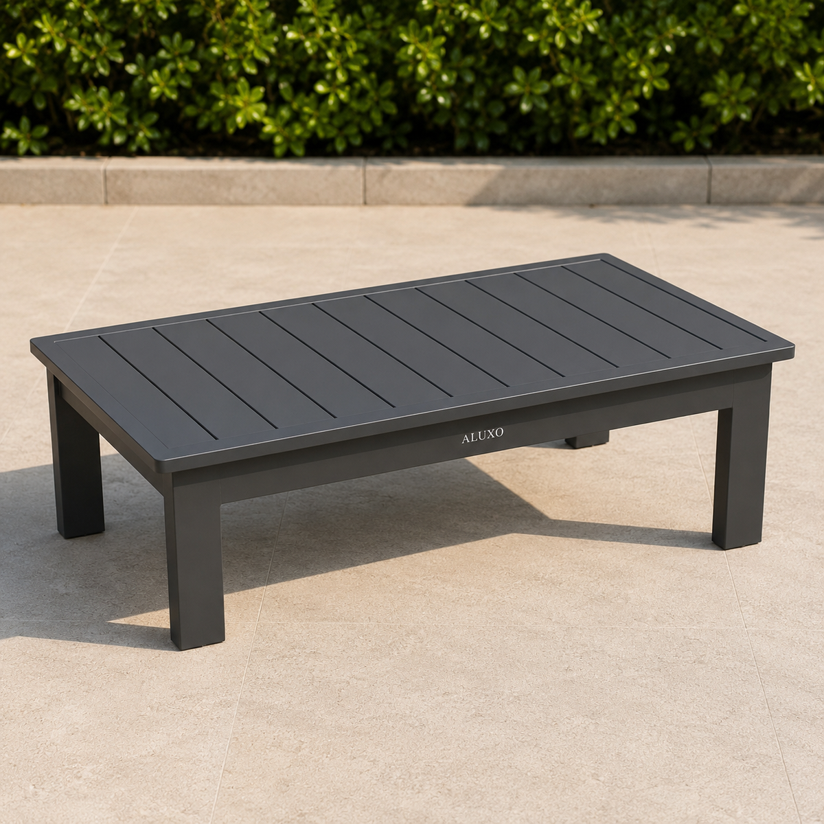 Dark grey matte metal outdoor coffee table on beige stone patio with green shrubs background