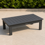 Dark grey matte metal outdoor coffee table on beige stone patio with green shrubs background