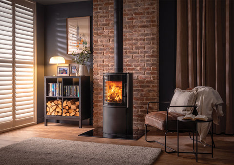 A modern Atlas wood burning stove in a stylish living room setting with a brick wall, wooden floor, and cozy furnishings.