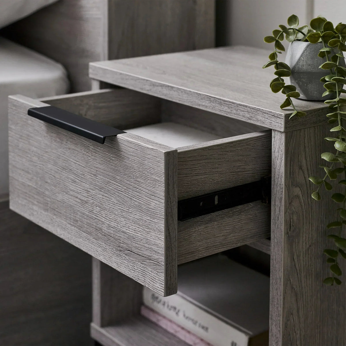 Grey oak bedside table with open drawer, black handle and potted plant on top