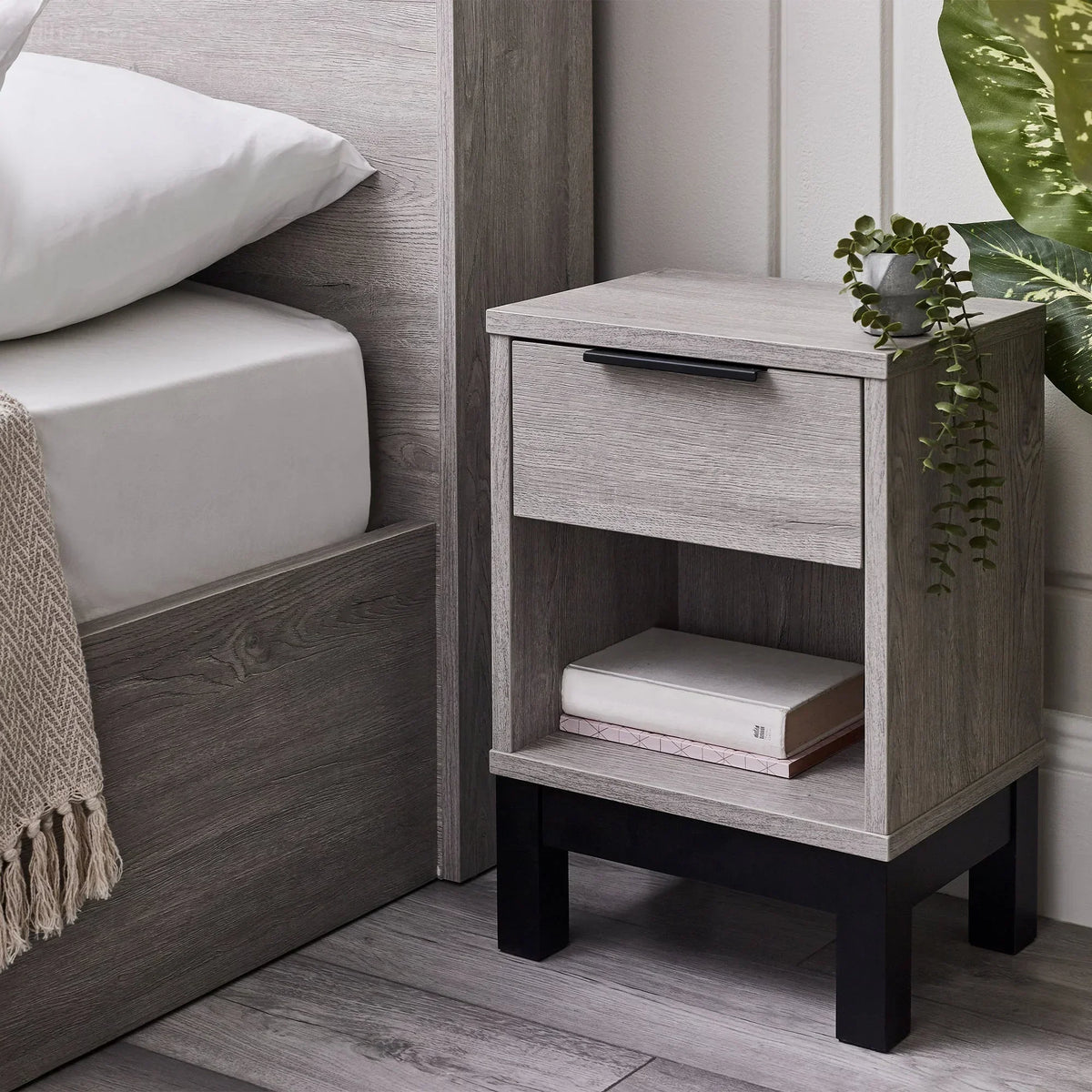 Grey oak 1 drawer bedside table with black legs beside bed, books and plant on top