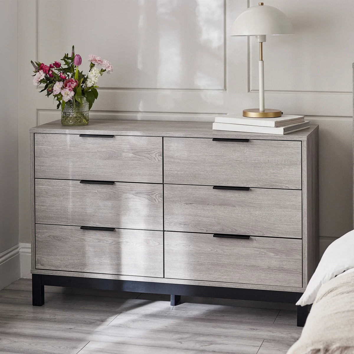 Light wood 6 drawer wide chest with black handles in a bedroom, styled with lamp and flowers