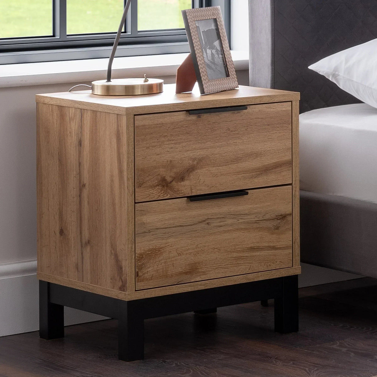 Oak two drawer bedside table with black handles beside bed and window
