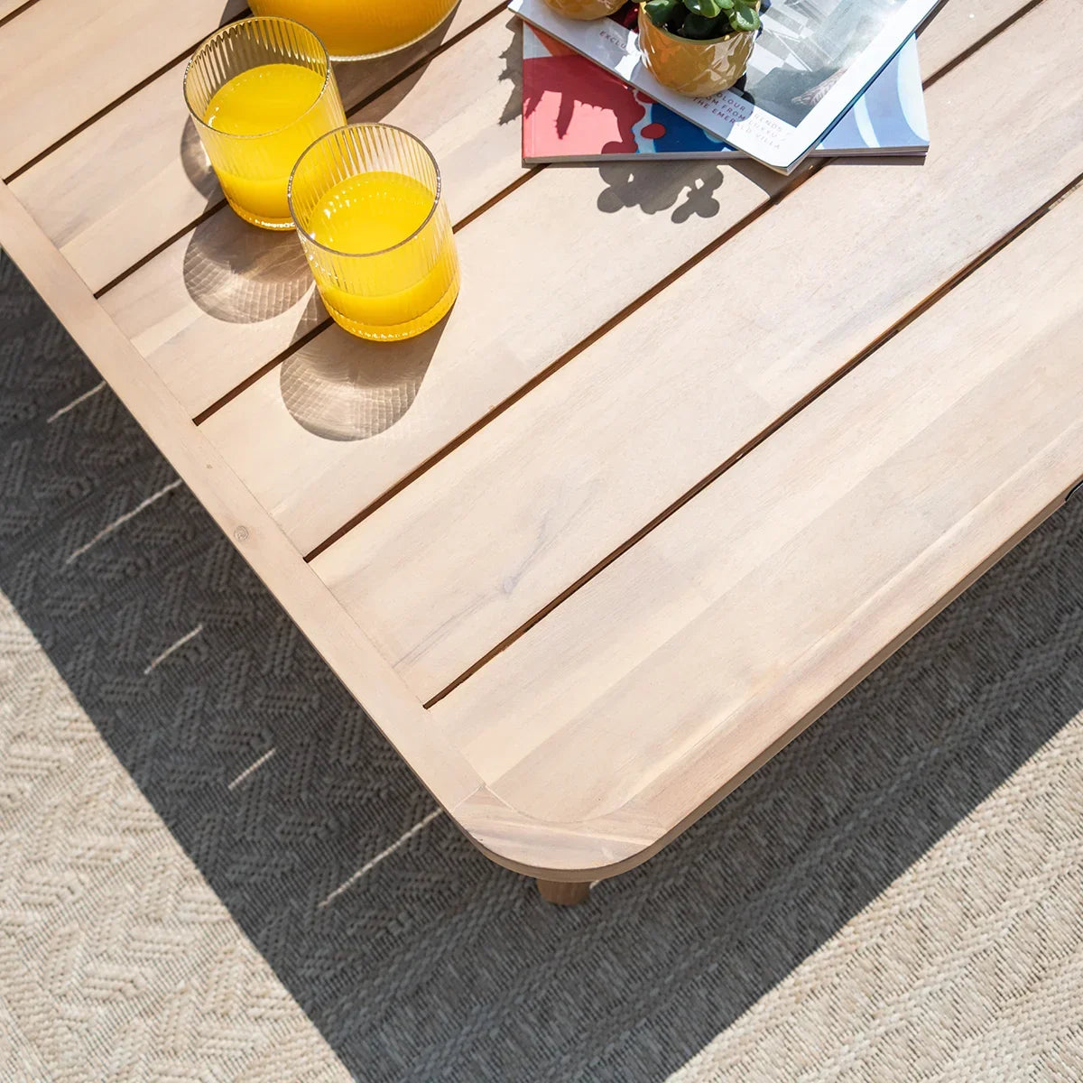 Light wood outdoor coffee table with drinks and magazines on textured patio rug