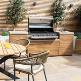 Outdoor wooden dining set with rattan chairs next to built-in gas barbecue and cabinets