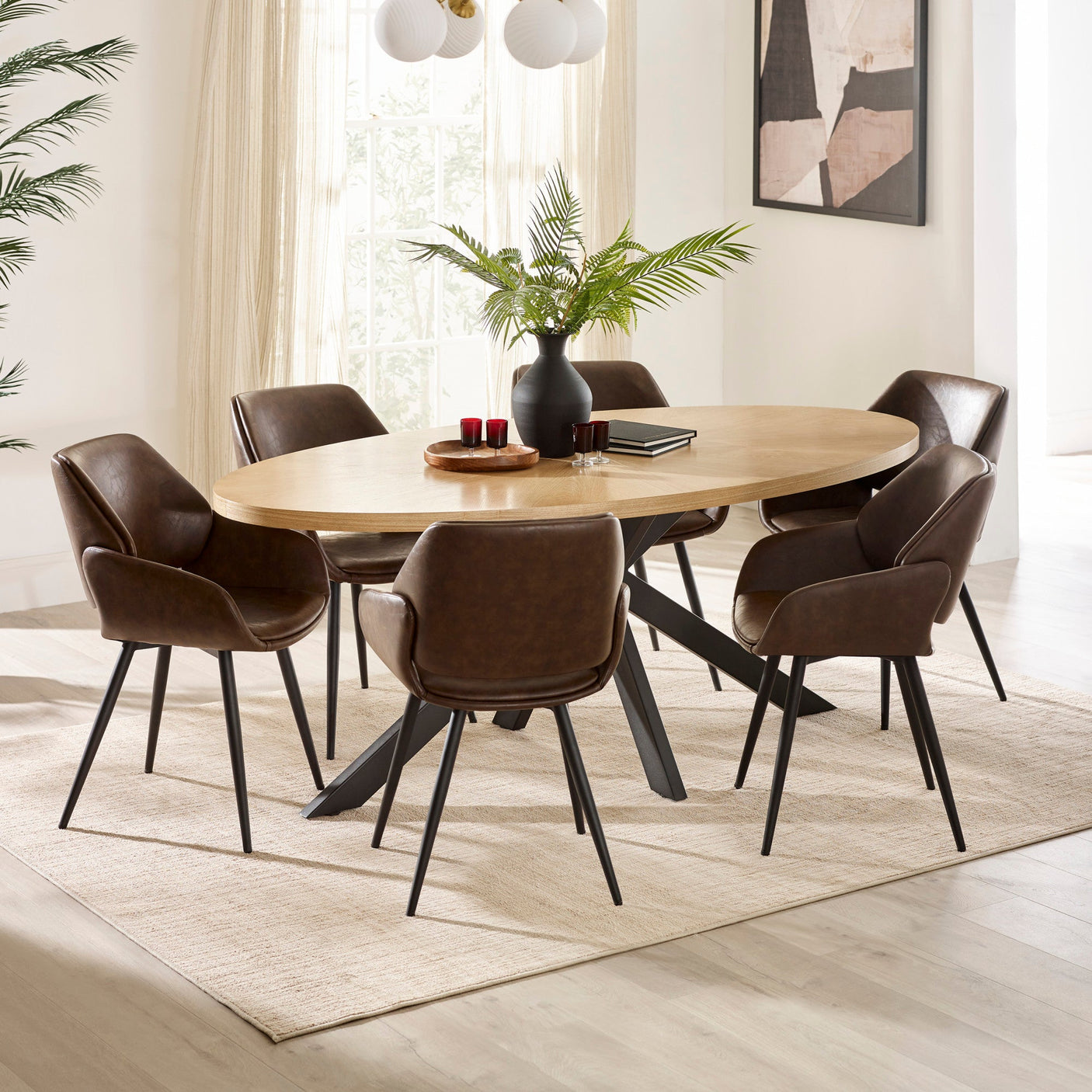 Light wood oval dining table with black legs on beige rug, six brown chairs, black vase with green foliage, and wall art