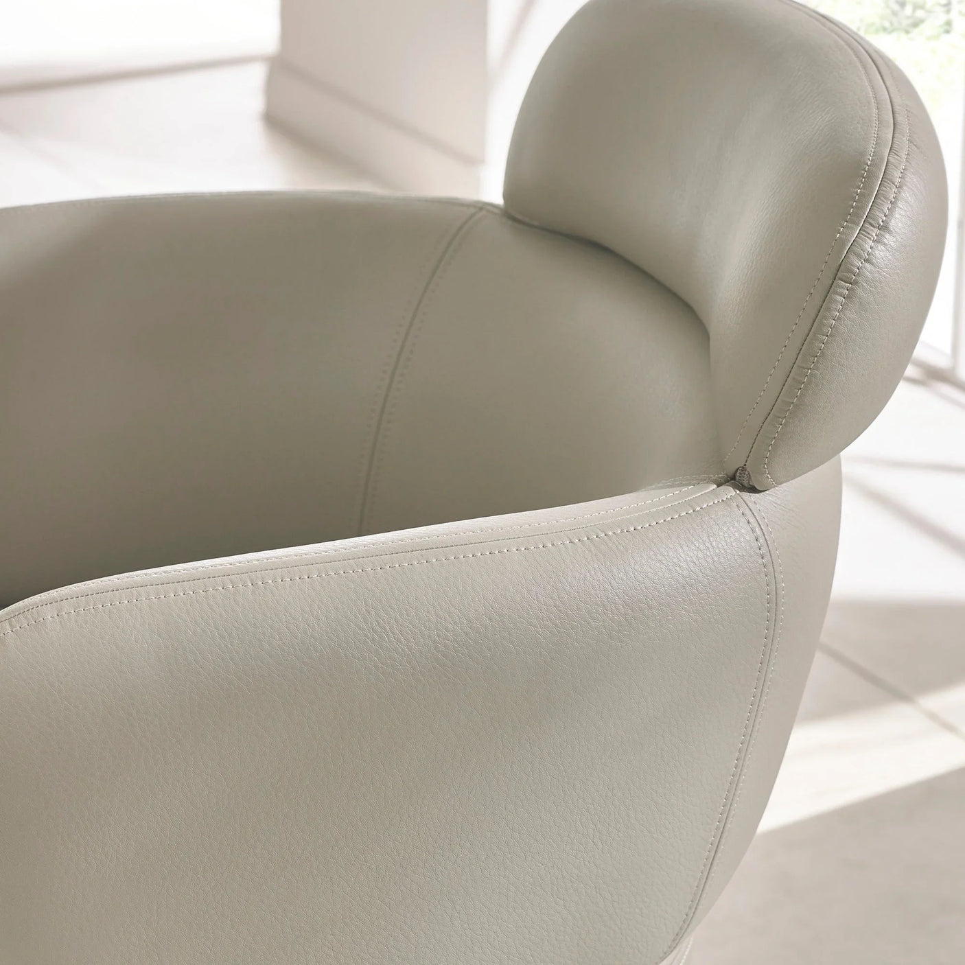 Light grey leather armchair with curved backrest and visible stitching in bright room