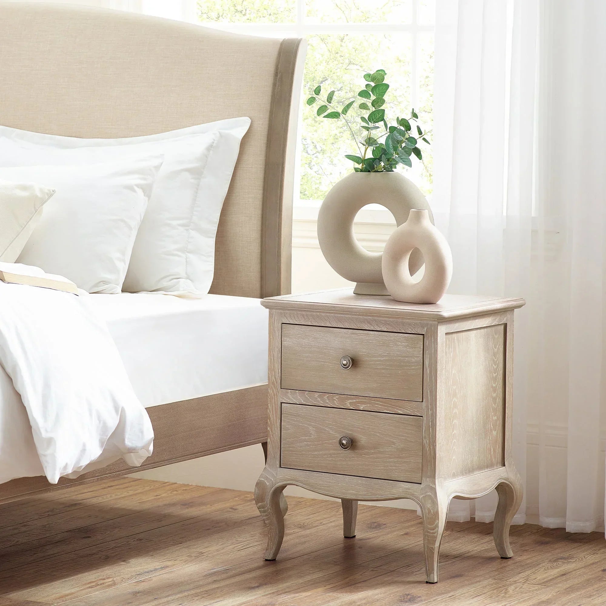 Light oak bedside table with two drawers beside bed, topped with modern beige vases