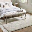Button tufted sitting bench in light beige fabric with carved wooden legs in a bedroom setting
