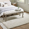 Button tufted sitting bench in light beige fabric with carved wooden legs in a bedroom setting